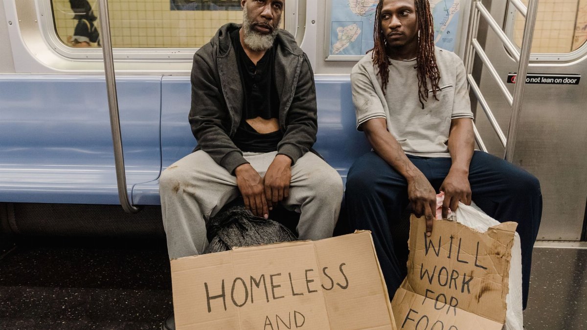 Two men sitting in a subway car holding signs conveying homelessness and hunger.
