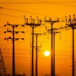 Silhouette of electric towers during sunset, highlighting power lines with a warm orange sky.