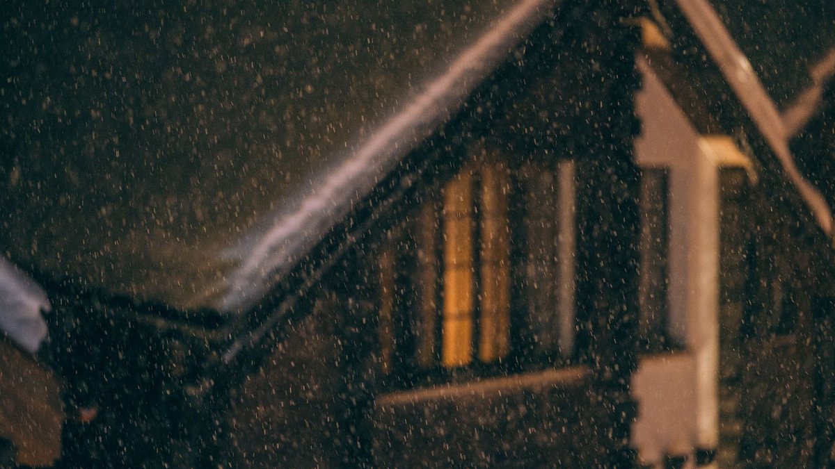 Exterior of small wooden cottage with snow on roof and burning electric lights in window during snowfall at cold winter night
