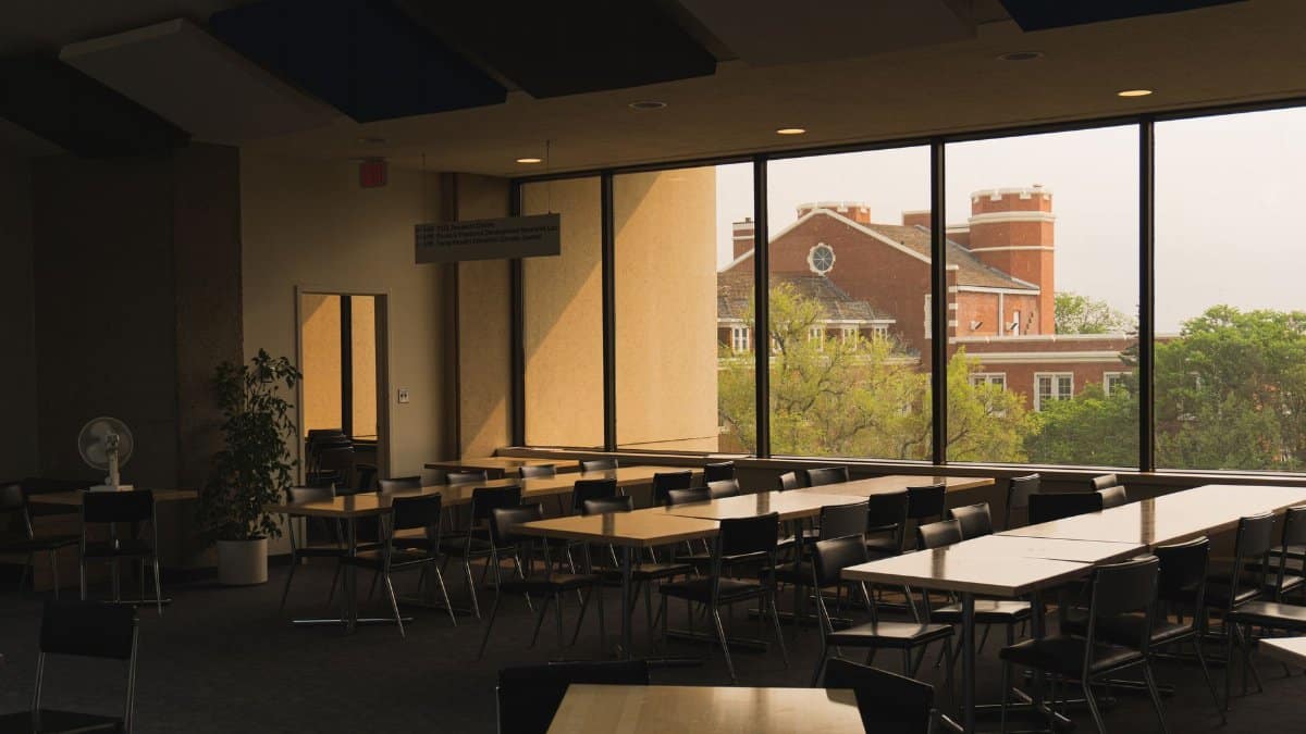 An empty university study hall with tables and chairs overlooking a campus building through large windows.