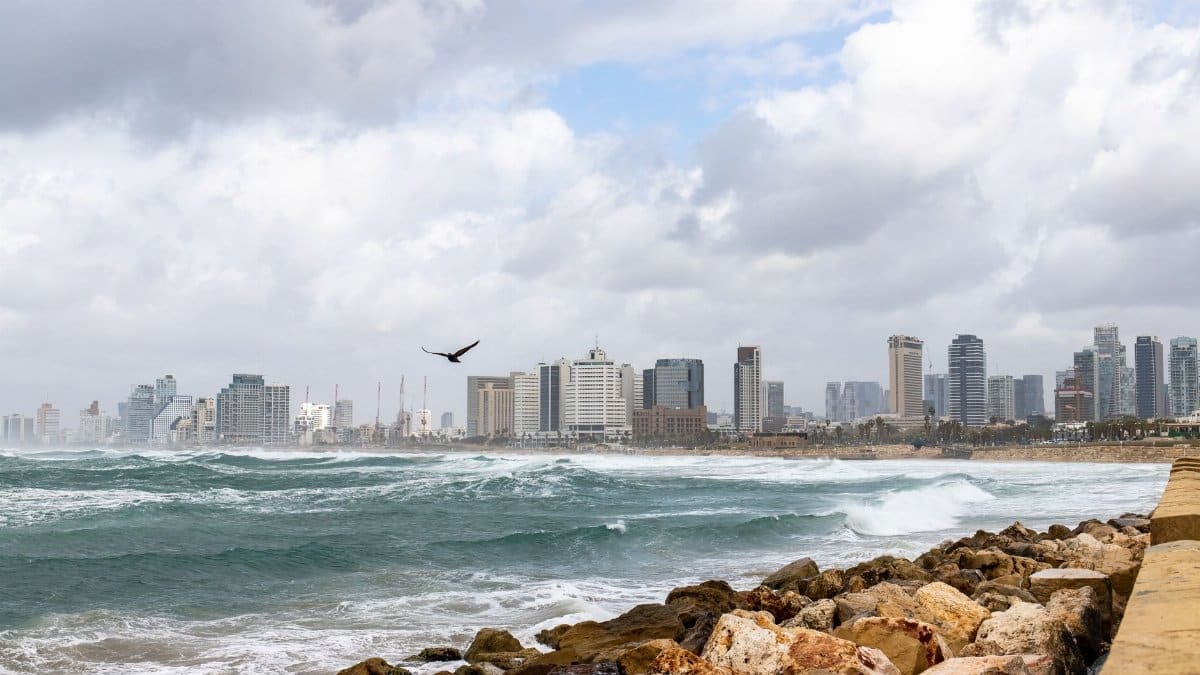Scenic view of Tel Aviv's skyline with rocky seashore and waves crashing, under a cloudy sky.