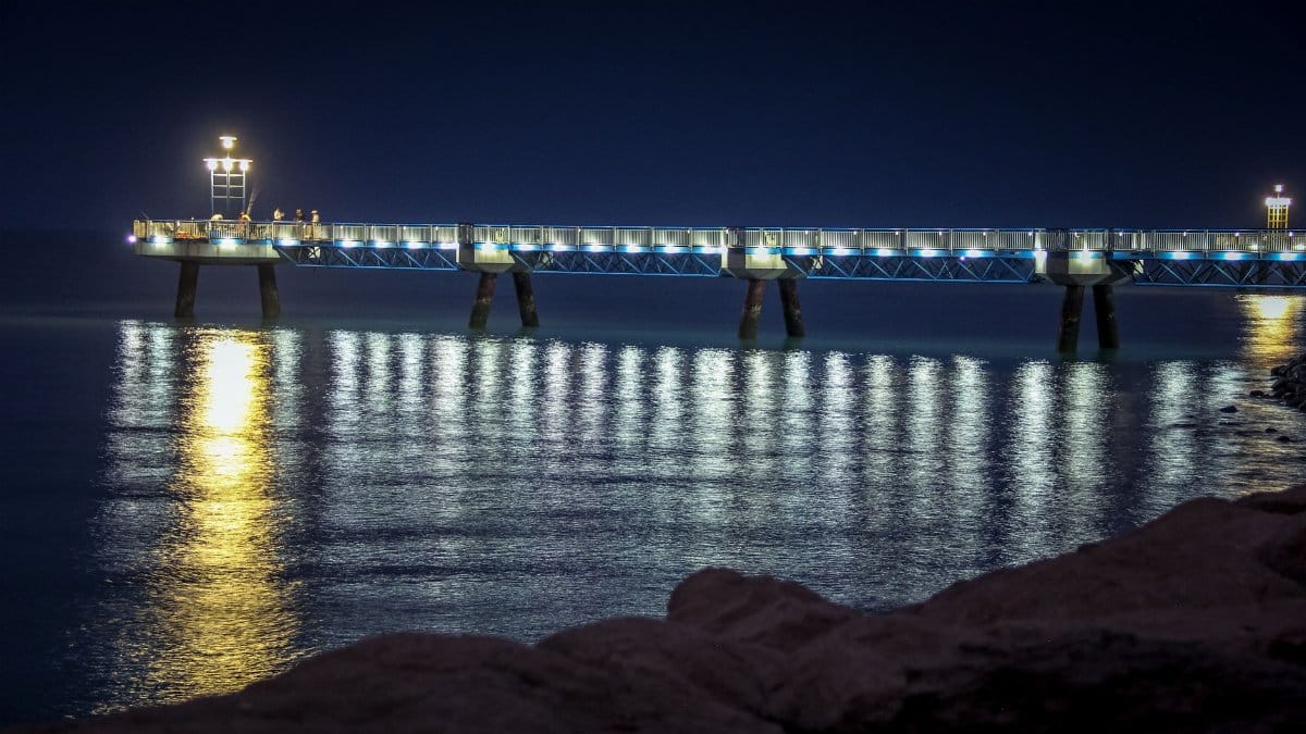 A serene night view of a brightly lit pier casting reflections on the calm ocean waters.