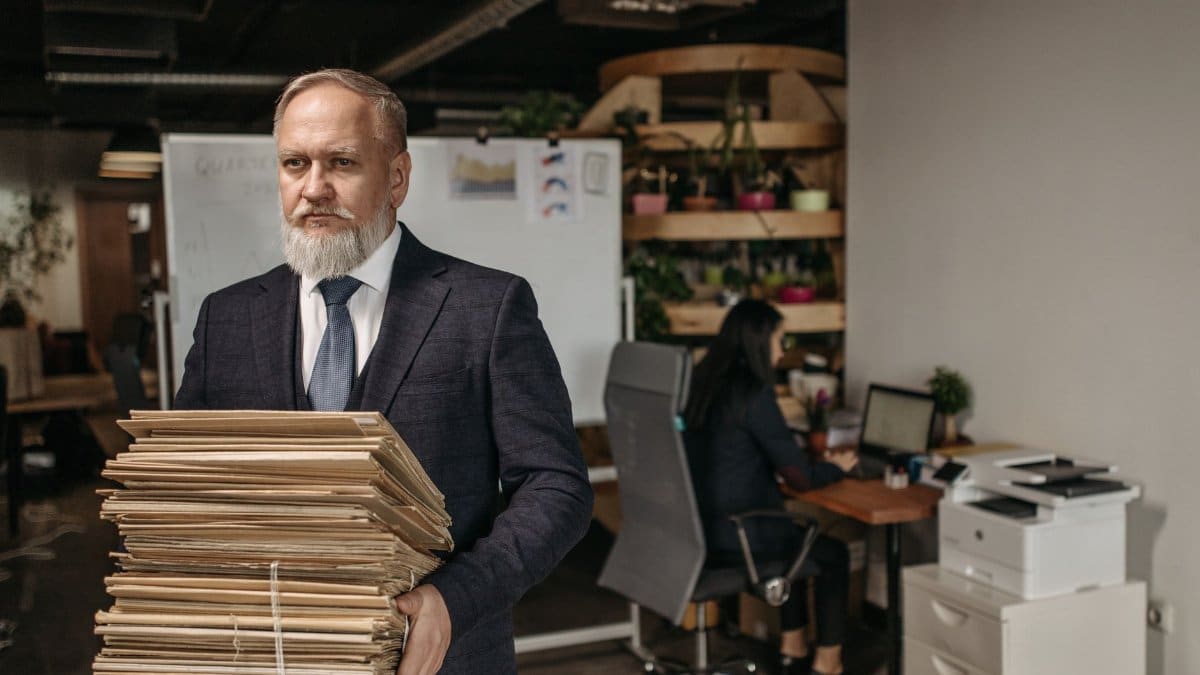 Elderly man in suit holding large pile of documents in a modern office setting.