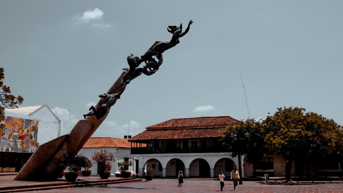 A striking sculpture dominates a sunny plaza with vibrant trees and architecture.