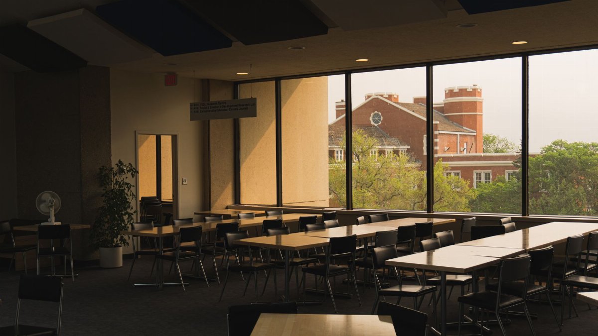 An empty university study hall with tables and chairs overlooking a campus building through large windows.
