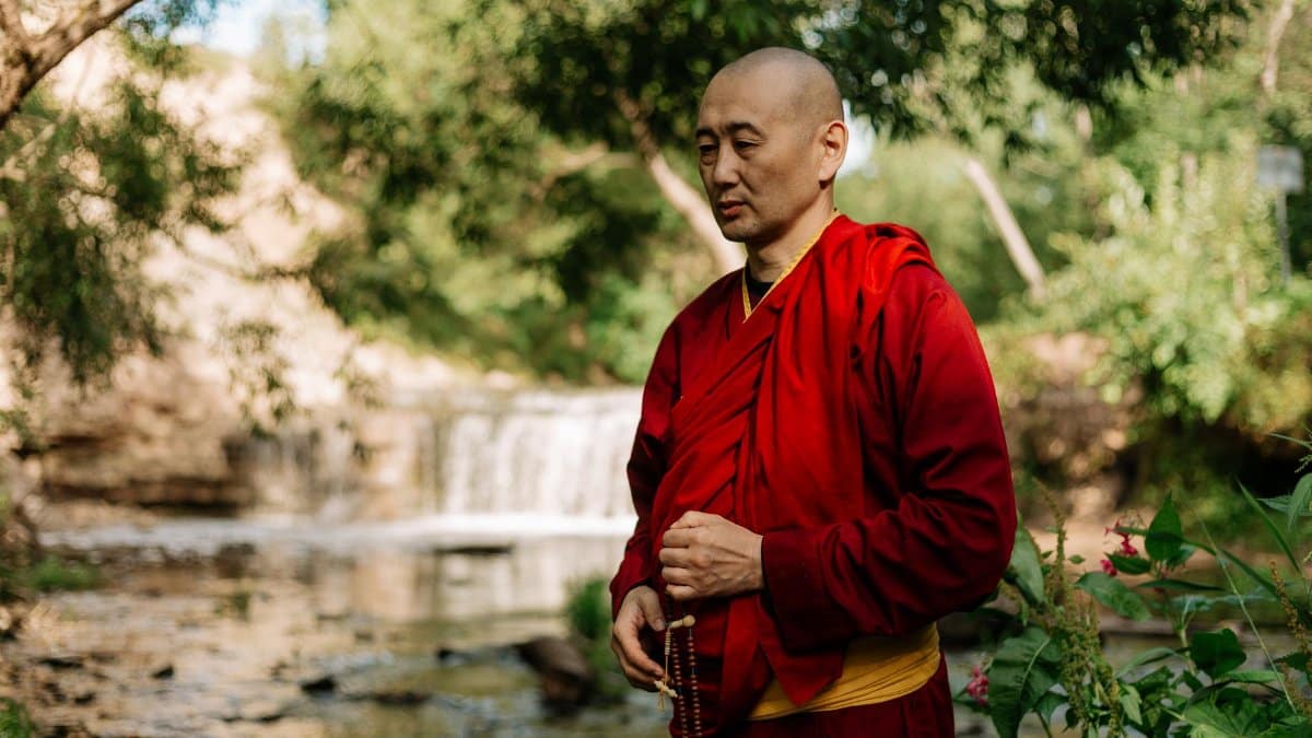 A serene Buddhist monk meditates by a stream in a lush green park, wearing traditional robes.
