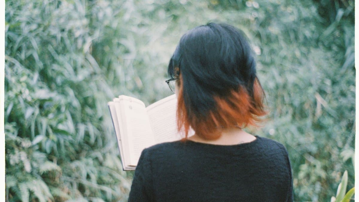A person with short hair reading a book outdoors, surrounded by lush greenery.