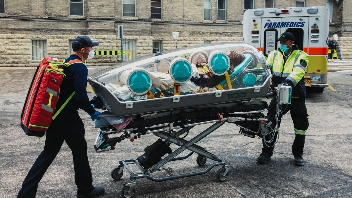 Emergency paramedics transport a patient in an isolation pod outside a medical facility.