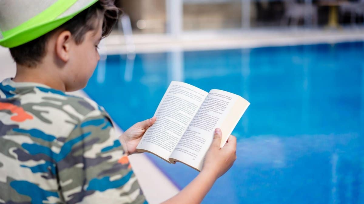 A young boy relaxes by the pool, reading a book in summer attire at a resort.
