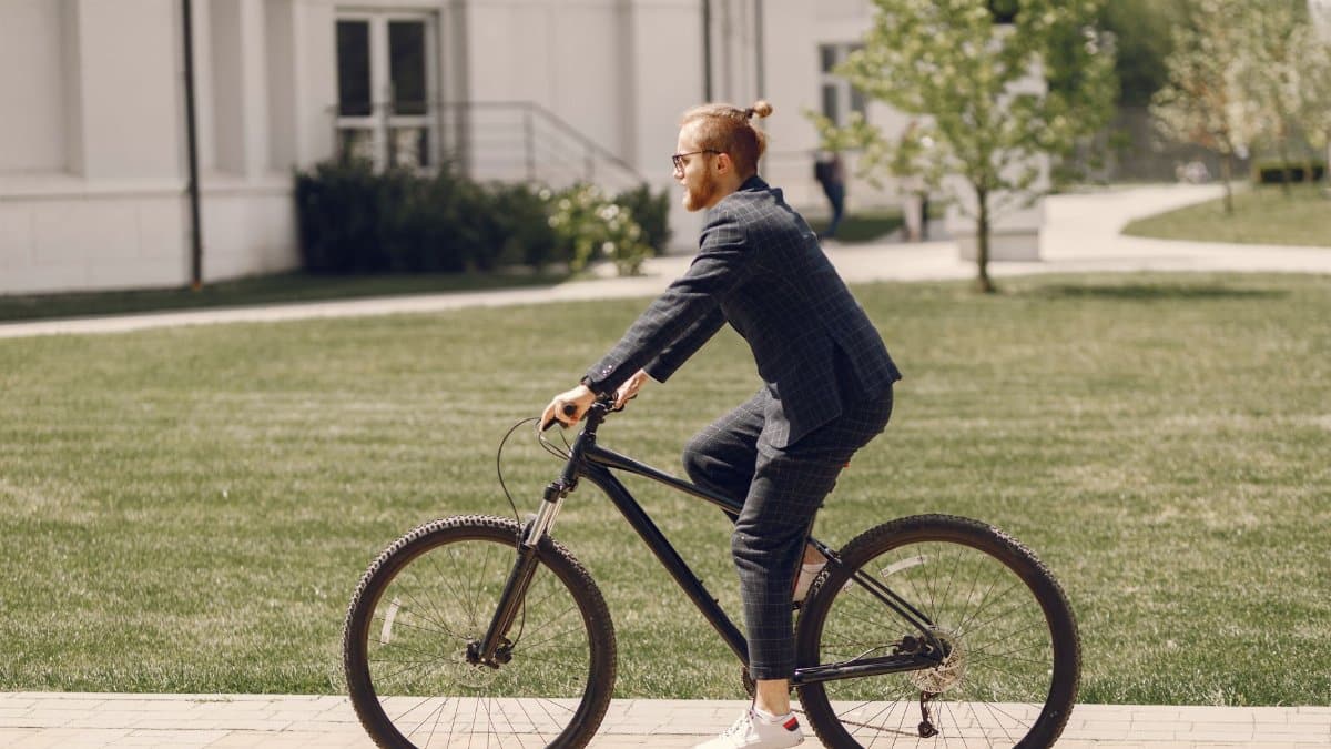 A stylish man in a suit confidently rides a bicycle through a green park on a sunny day.