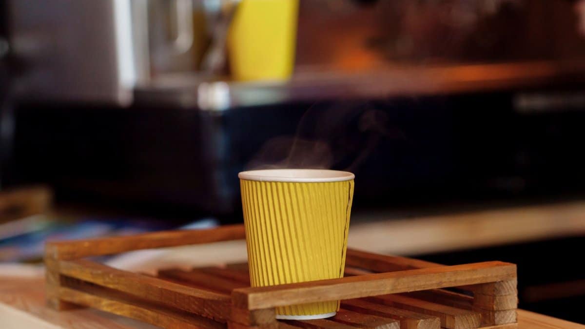 Close-up of a steaming yellow paper cup on a wooden tray in a cozy cafe setting.