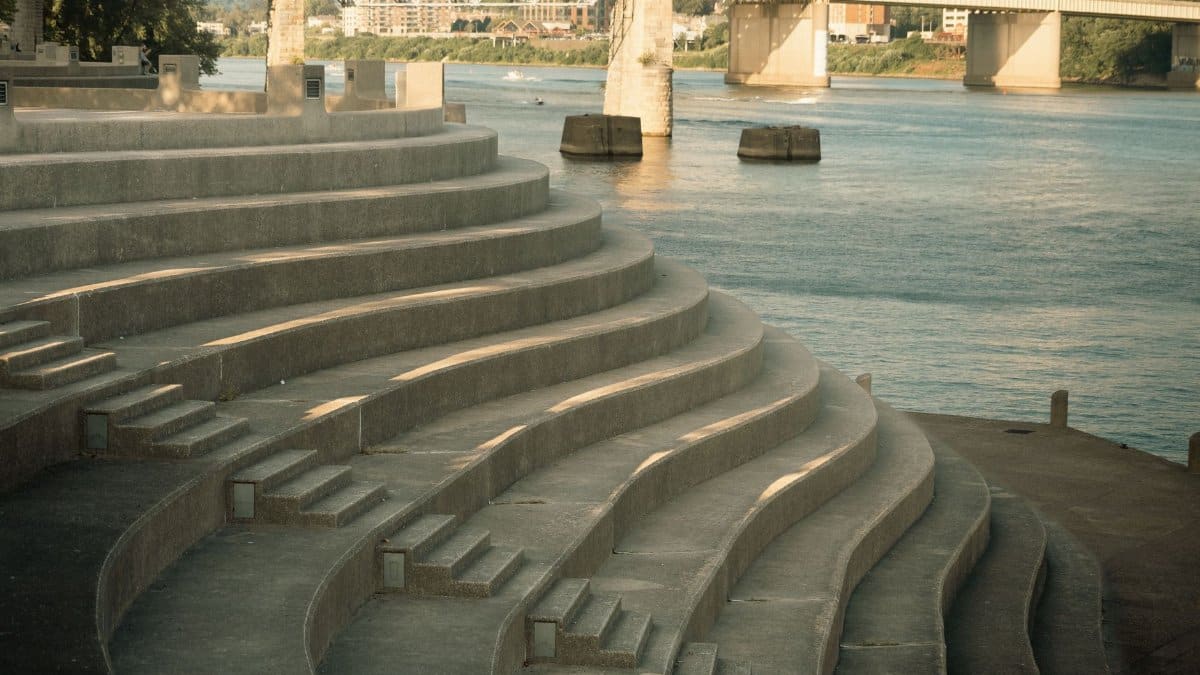 Concrete amphitheater steps beside a river with bridges in the background under daylight.