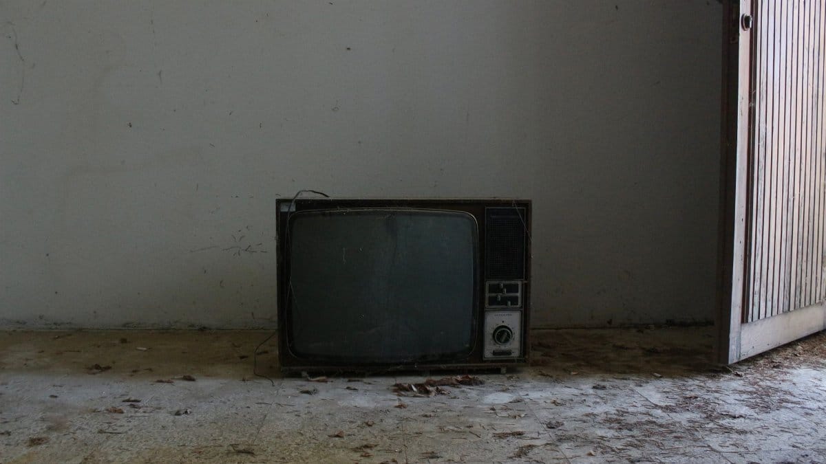 An old television set sits on a dusty floor in an abandoned room with a wooden door ajar.