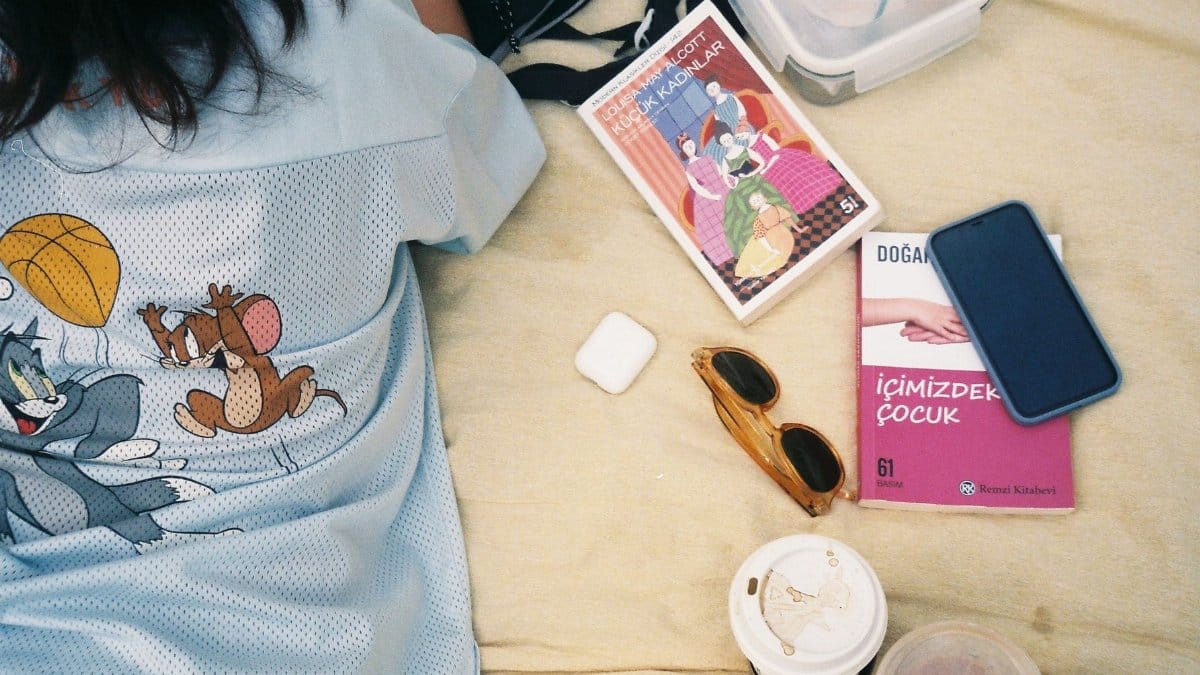 A person relaxes on a blanket with books, coffee, and a smartphone outdoors.