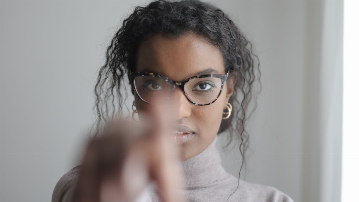 Confident African American female pointing with finger at camera looking seriously at camera through glasses on gray wall
