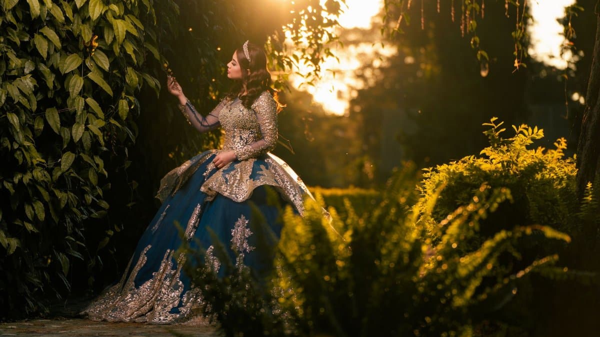 A woman in an elegant gown stands in a sunlit garden, evoking a fairy-tale atmosphere.