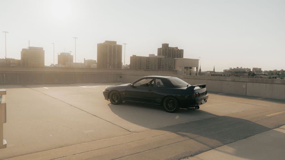 A Nissan Skyline parked in an urban rooftop lot with a cityscape and sunset in the background.