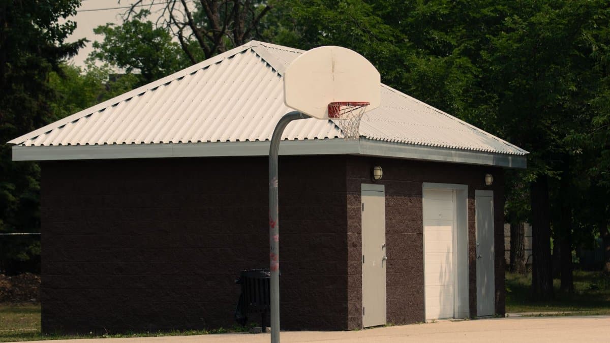 Basketball hoop at an outdoor court with a small building and trees surrounding it.