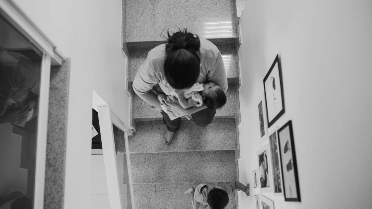 A mother carrying a baby descends a staircase with a young child in a modern home.