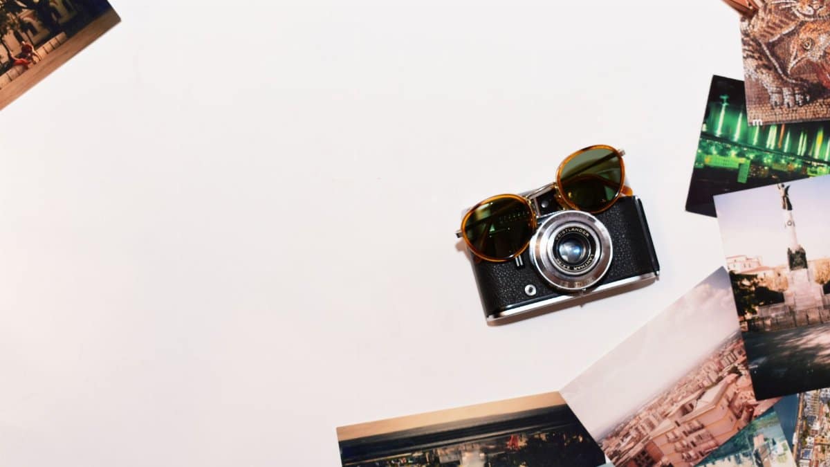 Top view of a vintage camera and sunglasses surrounded by travel photos on a white background.