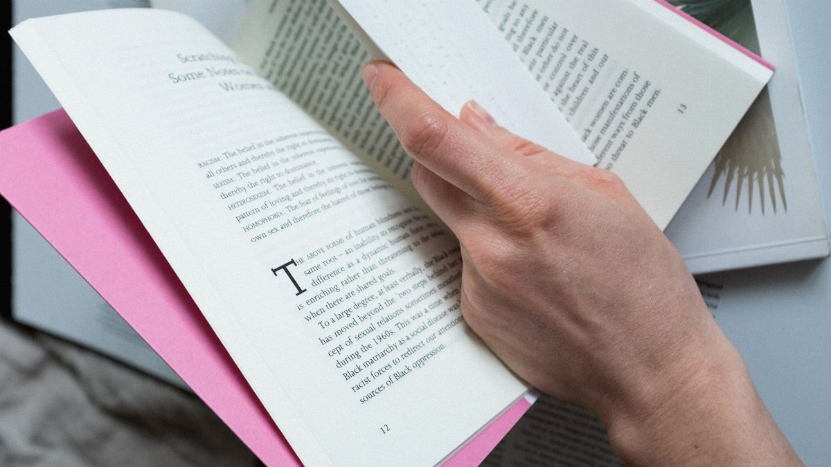 From above of crop person hand with paper reading book with pink cover on bed