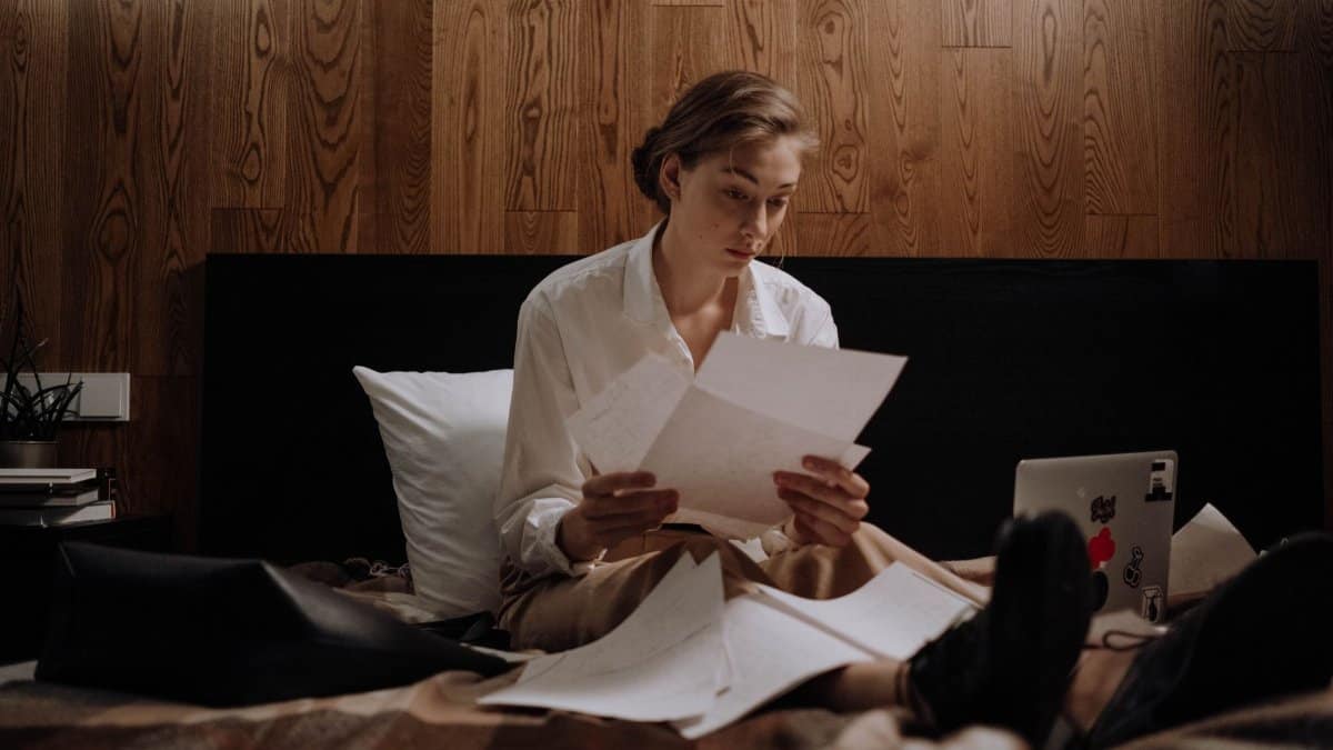 Woman sitting on bed, working from home with laptop and paperwork, focused and thoughtful.