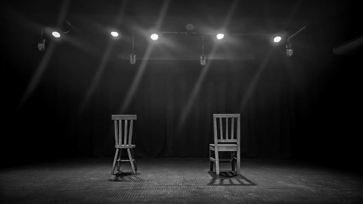 A dramatic black and white scene of two empty chairs spotlighted on a theater stage.