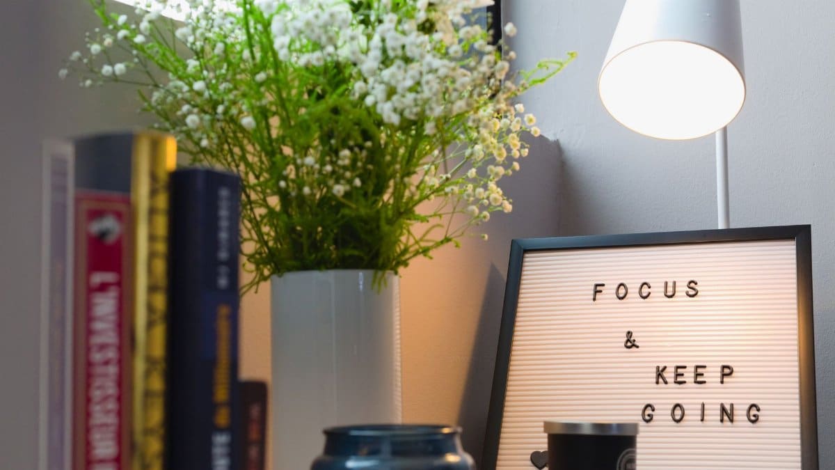 A cozy office space featuring books, a vase with flowers, lamp, and motivational sign.