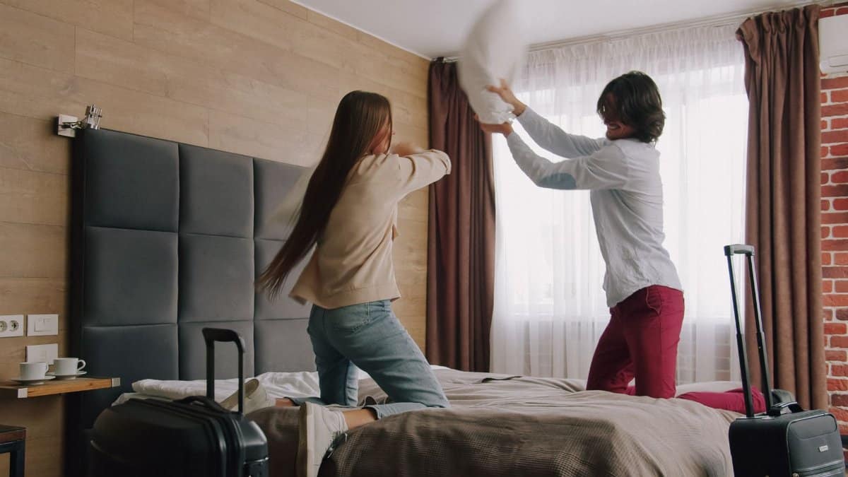 A young couple engaged in a playful pillow fight on a bed in a cozy hotel room.