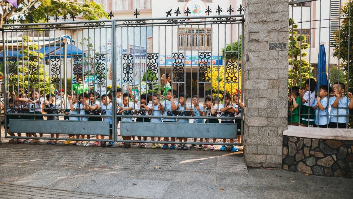 A group of young children, lined up and peering through a decorative school gate, enjoying a sunny day outdoors.