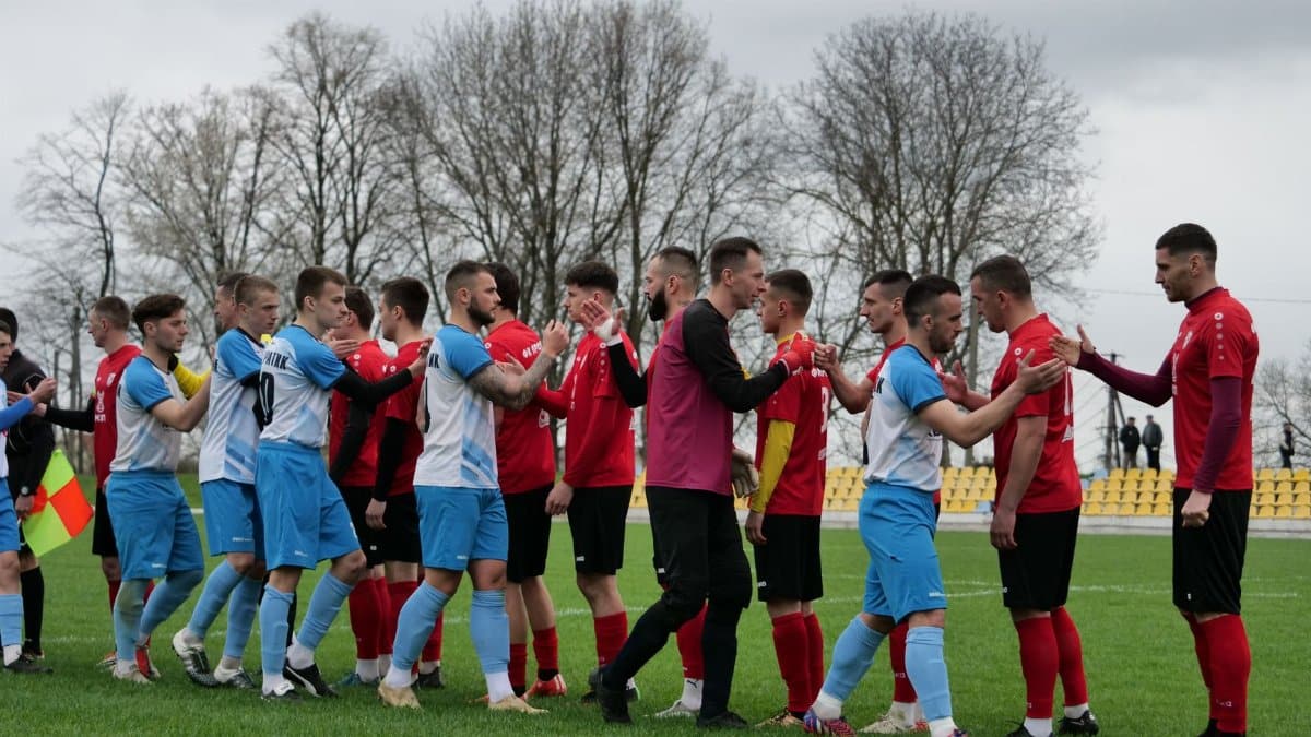 Football teams in red and blue kit shaking hands on a grassy pitch before the game.