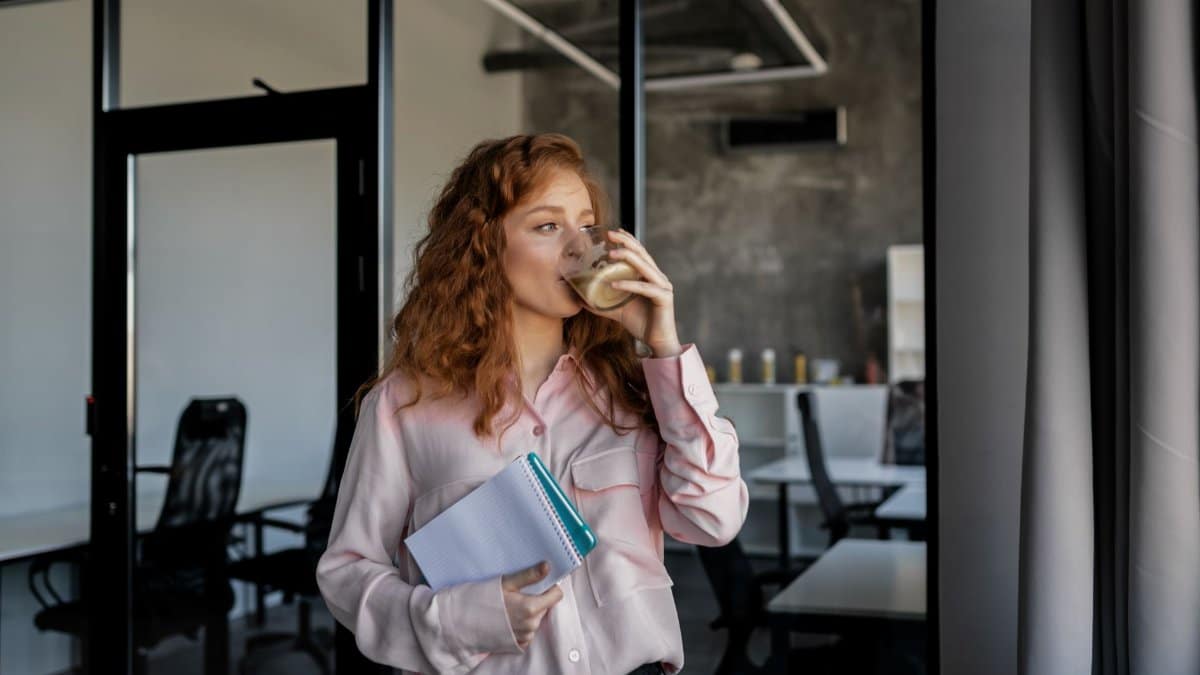 A redhead woman enjoys a coffee break while holding notebooks in a modern office.