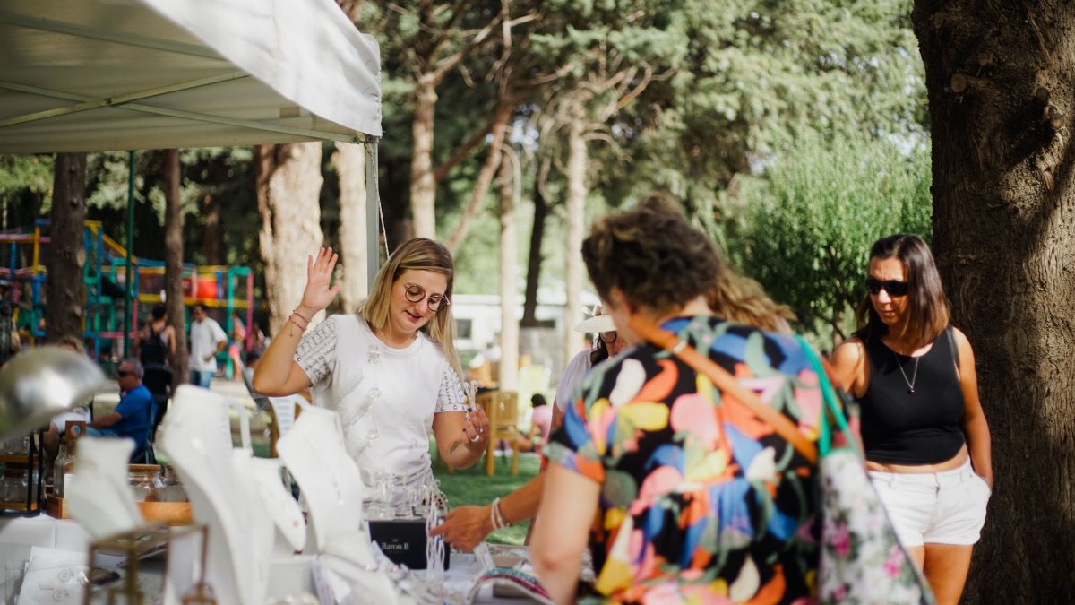 A lively scene of people shopping for jewelry at an outdoor market in a park during summer.