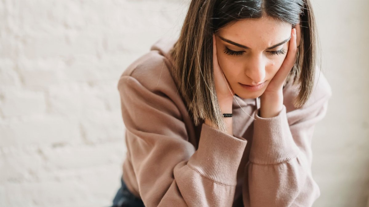 Crop unhappy female in casual wear touching cheeks and looking down while sitting near white wall in light room at home