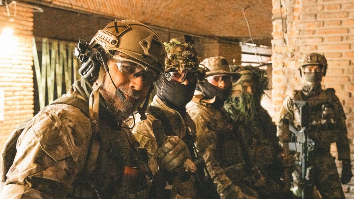 Team of soldiers in camouflage and helmets gathered indoors for a briefing.