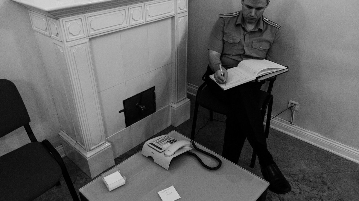 From above of concentrated male in police uniform sitting in room with tiled fireplace near coffee table with phone and holding hardcover notebook on knees and writing