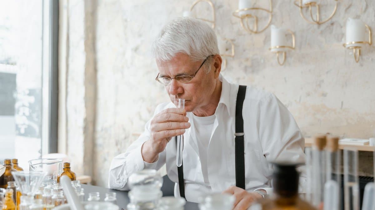 Senior man carefully smelling a test tube amidst laboratory glassware indicating a chemical experiment.