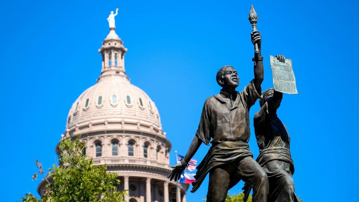 Statues with torch and document in front of Texas Capitol dome under clear blue sky in Austin.