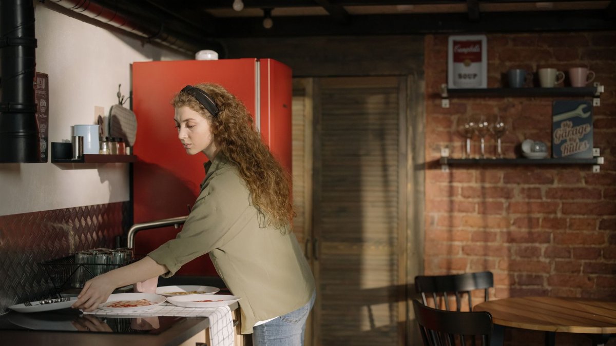 A woman arranging dinnerware in a cozy kitchen with a red fridge and brick wall.