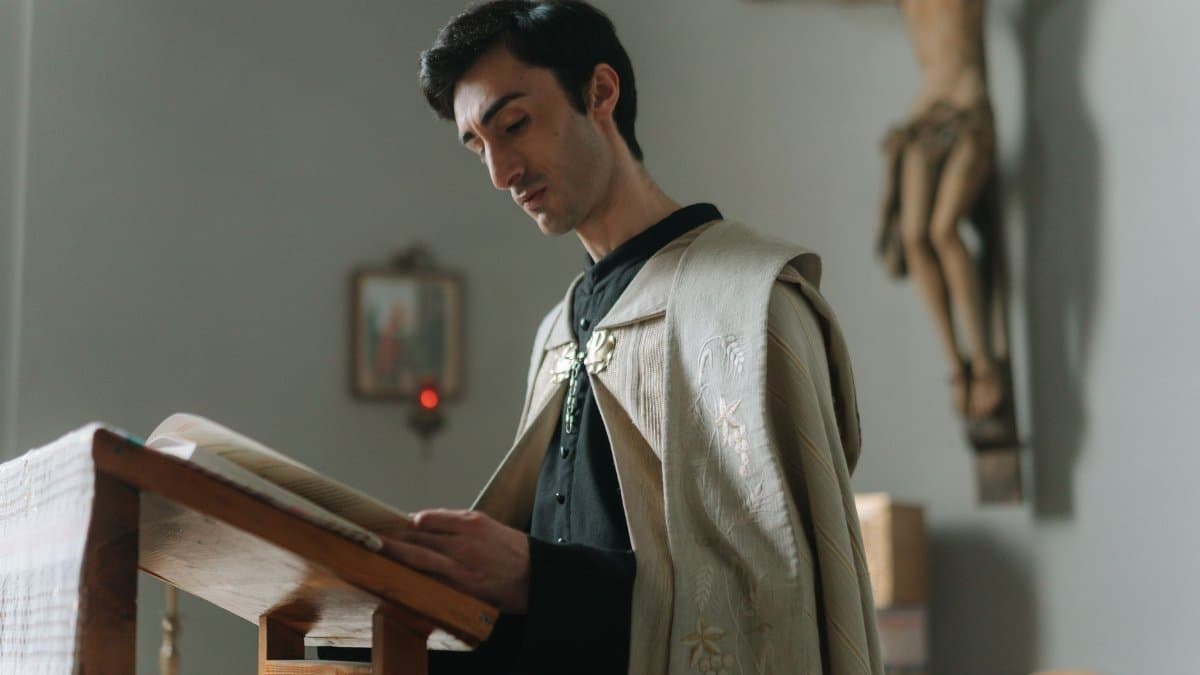 A priest reads from a book in a church, with a crucifix visible on the wall.