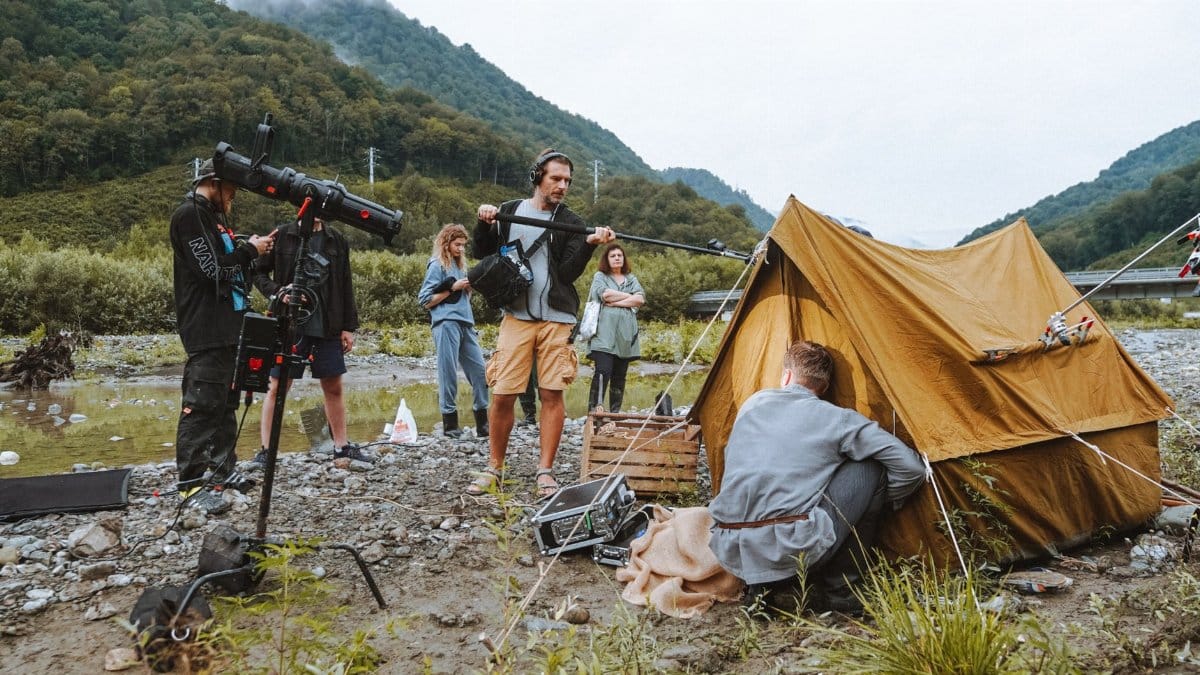 A film crew working with equipment and a tent by a river in a mountainous location.