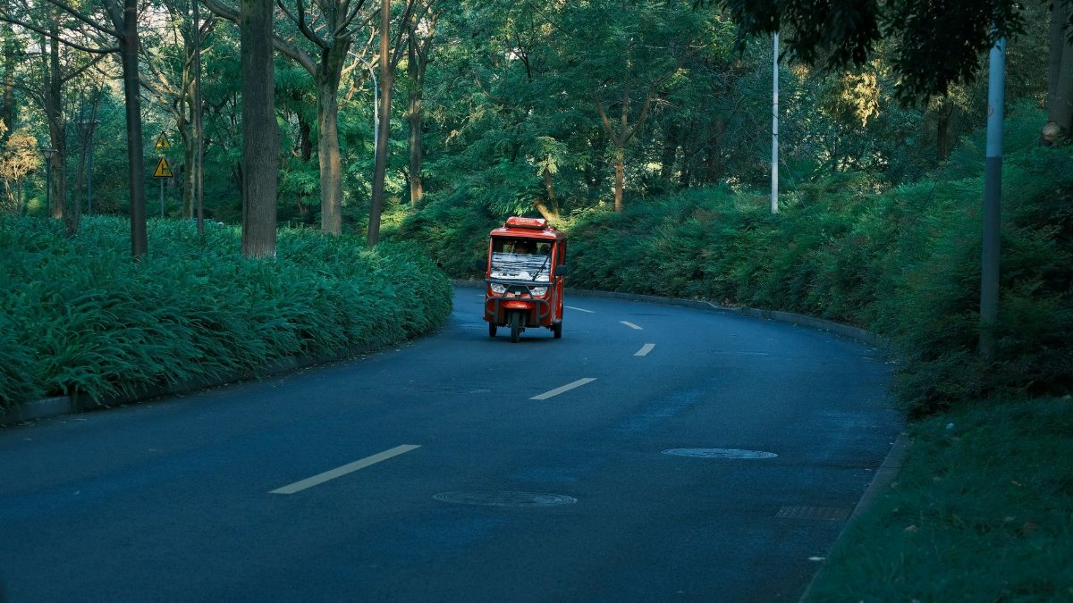 A bright red tuk-tuk driving on a peaceful, winding road surrounded by dense green forest.