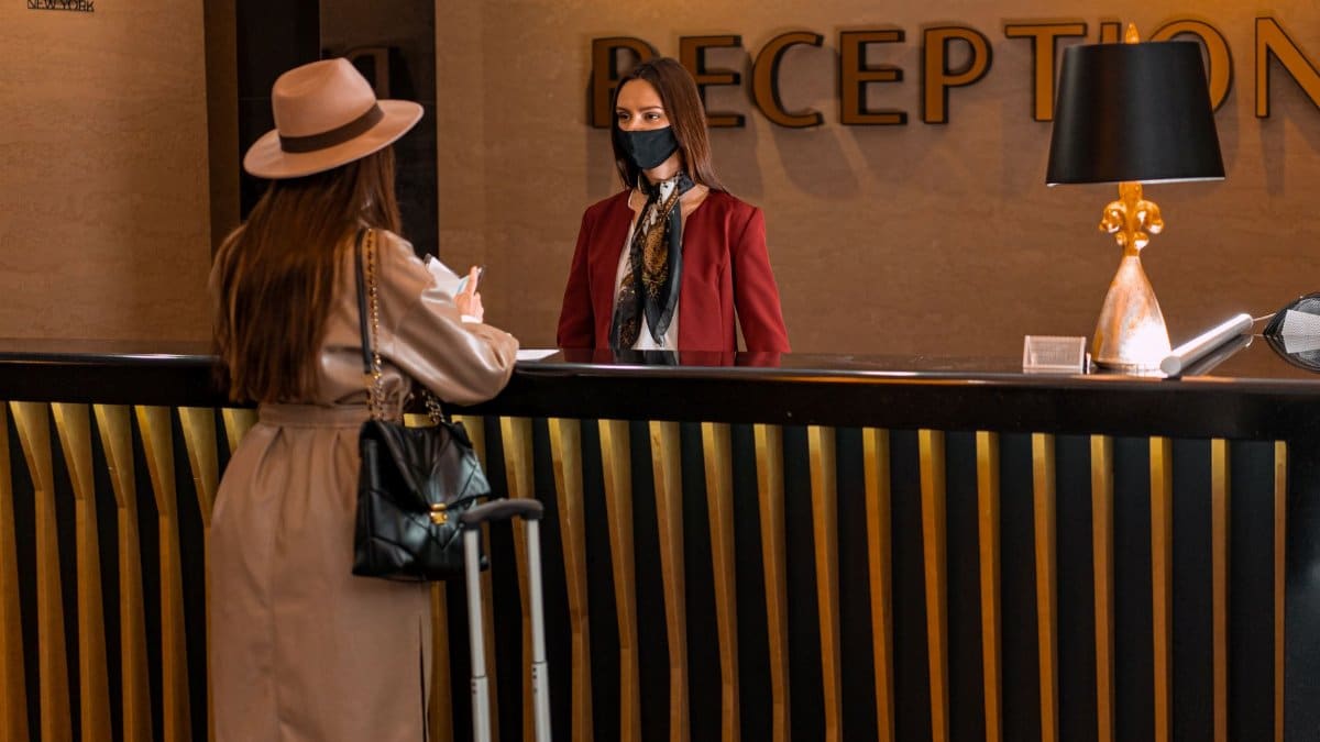 A woman speaks to a receptionist wearing a mask at a hotel reception.