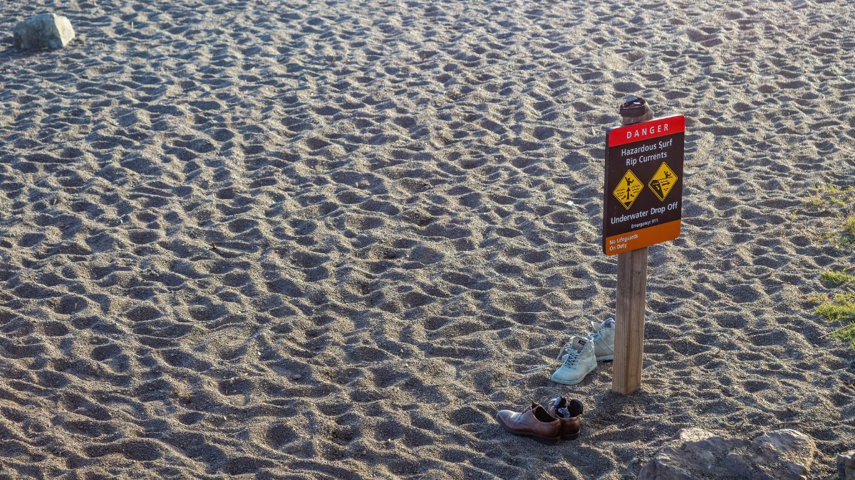 A beach warning sign stands on the sandy shore with two pairs of shoes nearby, emphasizing caution.