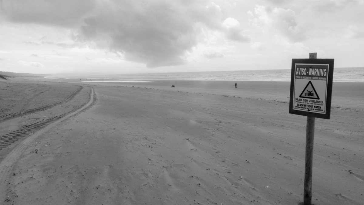 Monochrome beach view with a warning sign and dramatic clouds overhead.