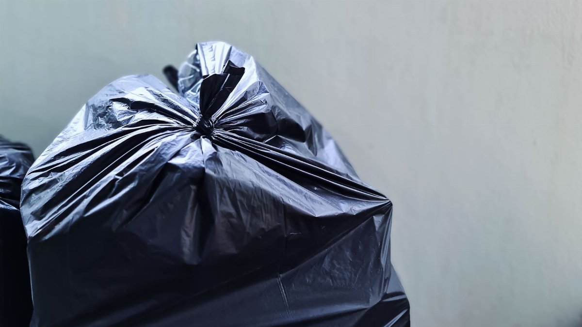 A close-up photograph of a black plastic garbage bag against a plain concrete wall, highlighting waste management.