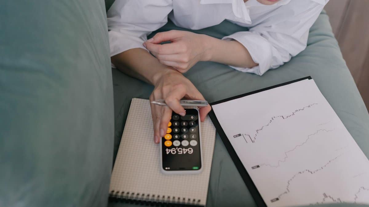 A woman works indoors using a smartphone calculator and analyzing financial graphs, symbolizing modern finance.
