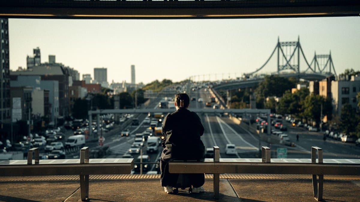A person sits on a bench watching traffic and a bridge in New York City during daytime.
