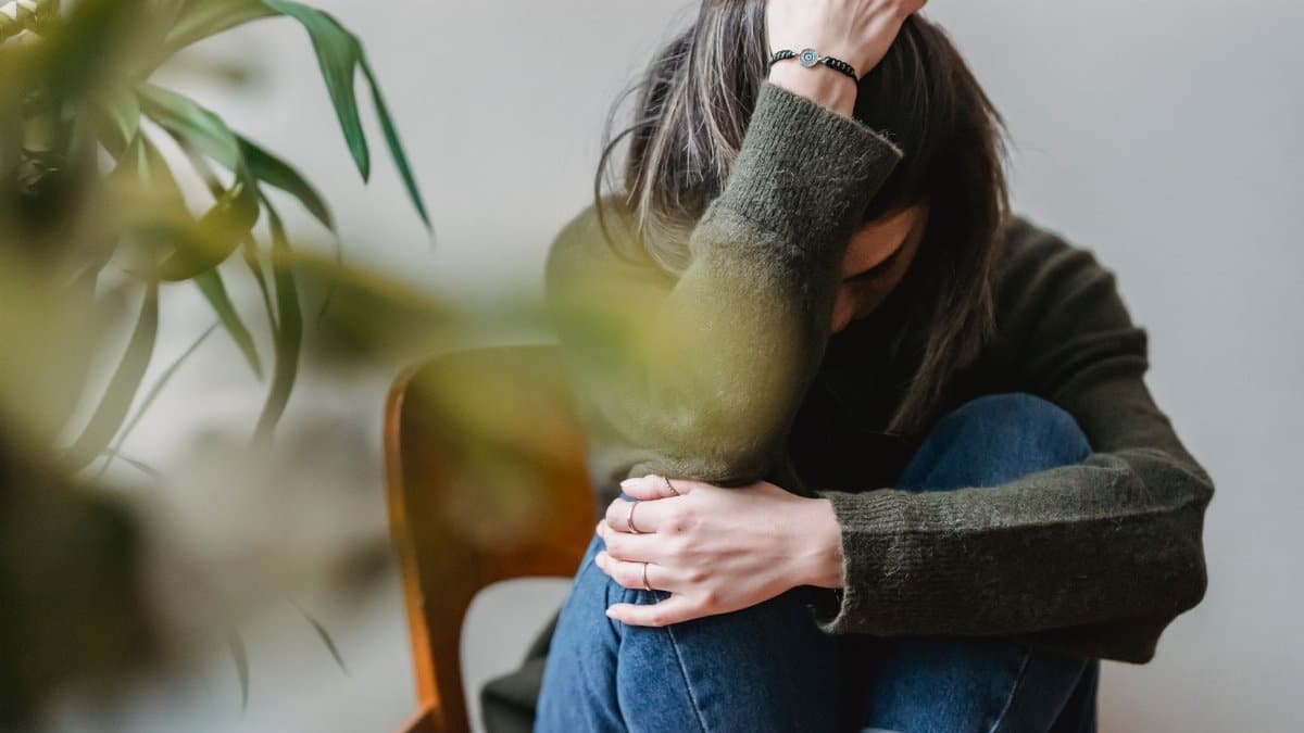 Anonymous young sorrowed female in casual outfit touching dark hair and embracing knees while sitting on chair at home