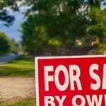 Close-up of a red 'For Sale By Owner' sign alongside a rural road with trees.